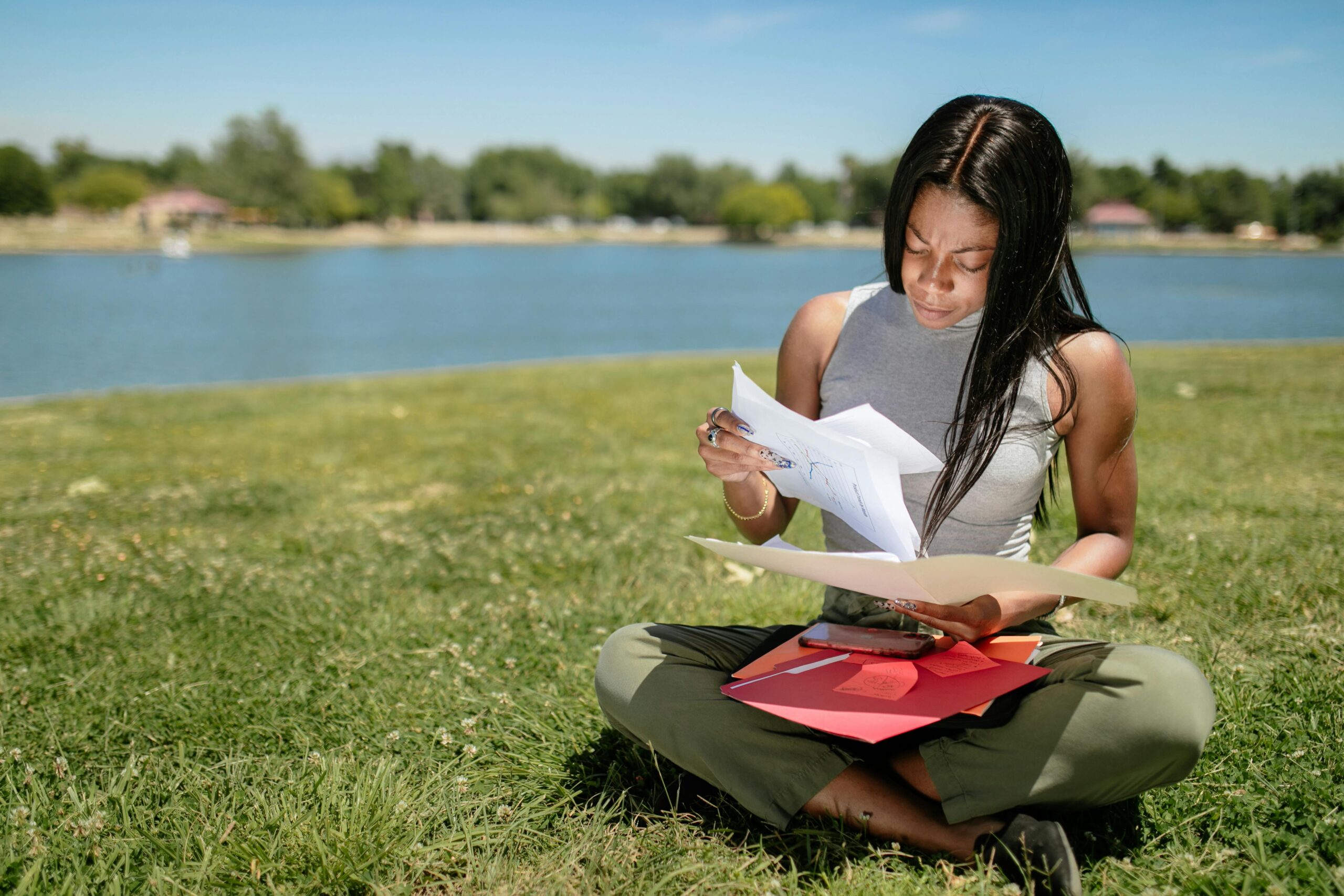 A woman sits on grass by a lake, examining papers on a sunny day.